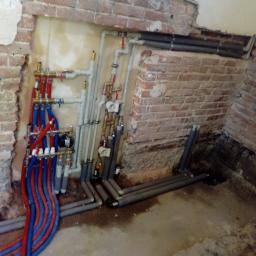 Edward Wiśniewski - Rozdzielacz instalacji centralnego ogrzewania z czerwono-niebieskimi rurami, zamontowany na tle surowej ściany z cegły z widocznymi śladami po kuciu, z szarymi rurami biegnącymi wzdłuż ściany...