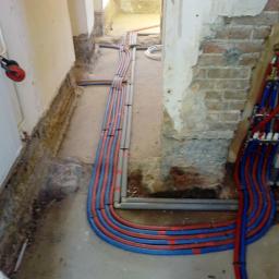 Edward Wiśniewski - Nowe rury instalacji grzewczej i hydraulicznej ułożone wzdłuż ściany, widoczne zawory i przyłącza, fragment odsłoniętej cegły, w tle dalsza część instalacji.