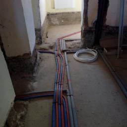 Edward Wiśniewski - Wnętrze w trakcie remontu z odsłoniętymi instalacjami: rury grzewcze i wodociągowe w kolorach niebieskim, czerwonym i szarym, prowadzone wzdłuż ściany i podłogi. Fragment ściany z odsłoniętą cegłą...