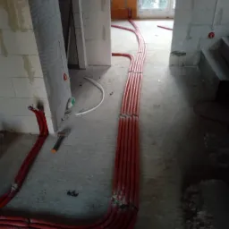 Układanie wiązki czerwonych rur instalacji grzewczej w surowym wnętrzu budynku, widoczne bloczki betonowe i fragment okna.