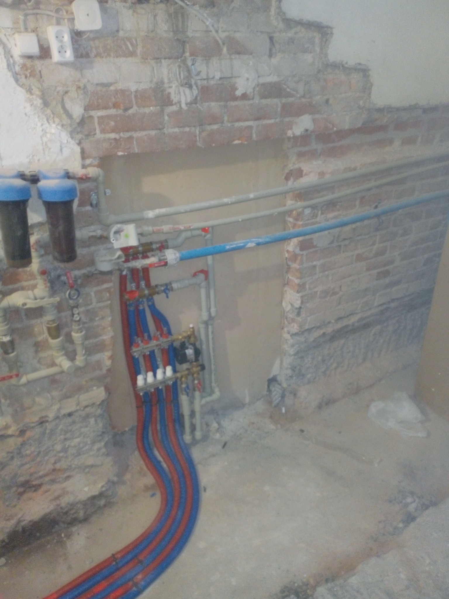Wnętrze w trakcie prac instalacyjnych: widoczny rozdzielacz z czerwono-niebieskimi rurami, filtry wody, oraz odsłonięta ceglana ściana z fragmentami tynku i przewodami elektrycznymi.