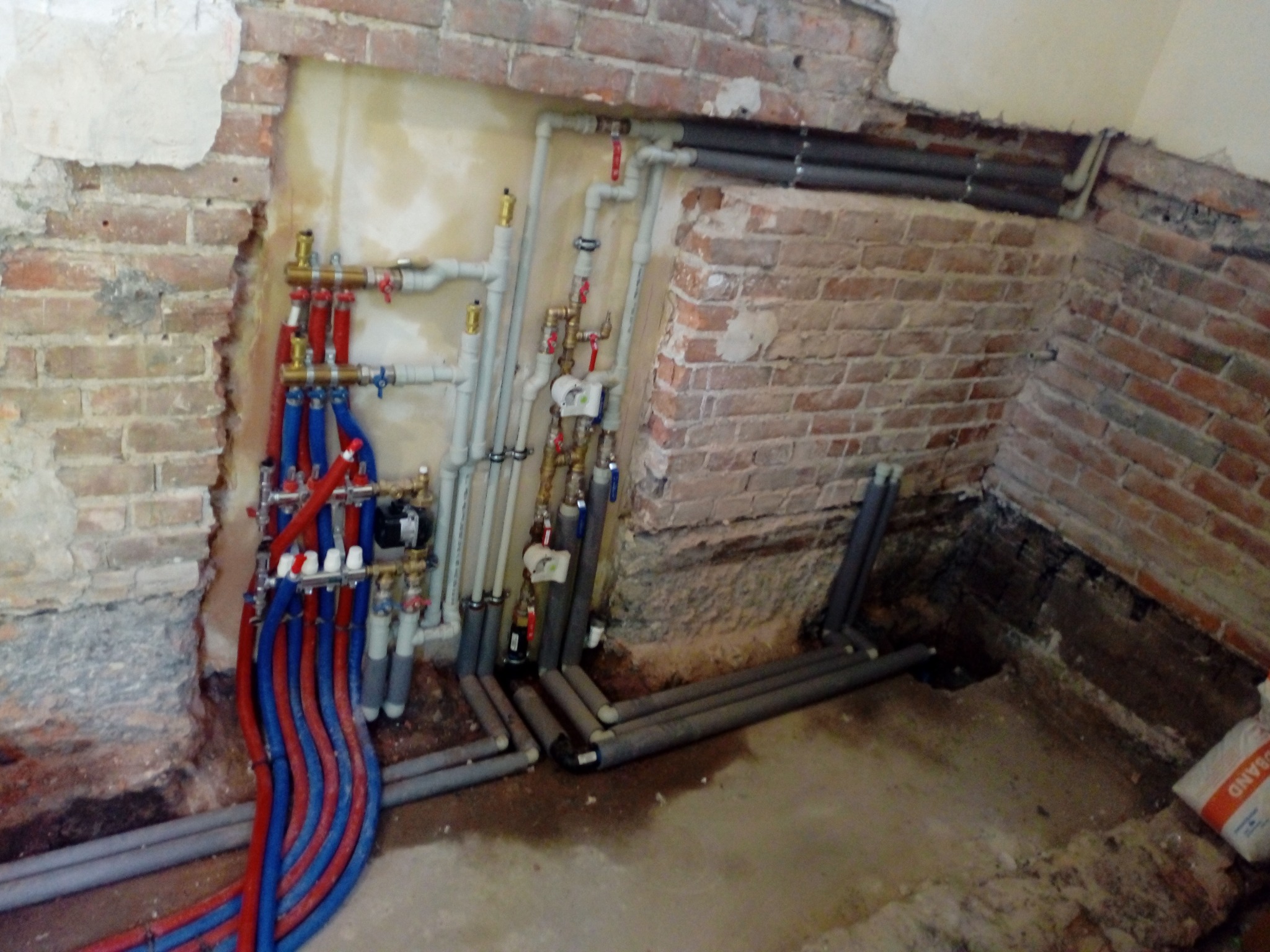 Rozdzielacz instalacji centralnego ogrzewania z czerwono-niebieskimi rurami, zamontowany na tle surowej ściany z cegły z widocznymi śladami po kuciu, z szarymi rurami biegnącymi wzdłuż ściany...