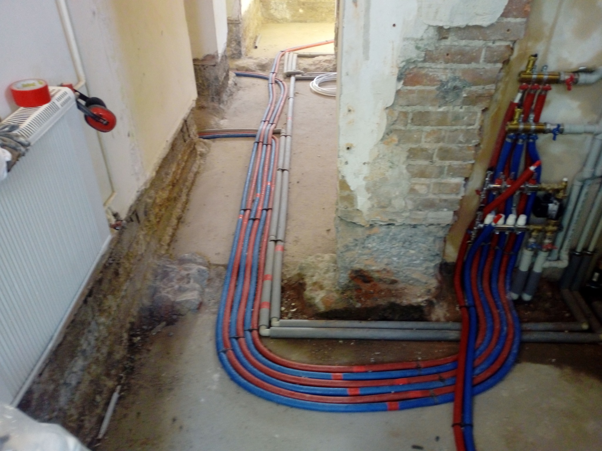 Nowe rury instalacji grzewczej i hydraulicznej ułożone wzdłuż ściany, widoczne zawory i przyłącza, fragment odsłoniętej cegły, w tle dalsza część instalacji.