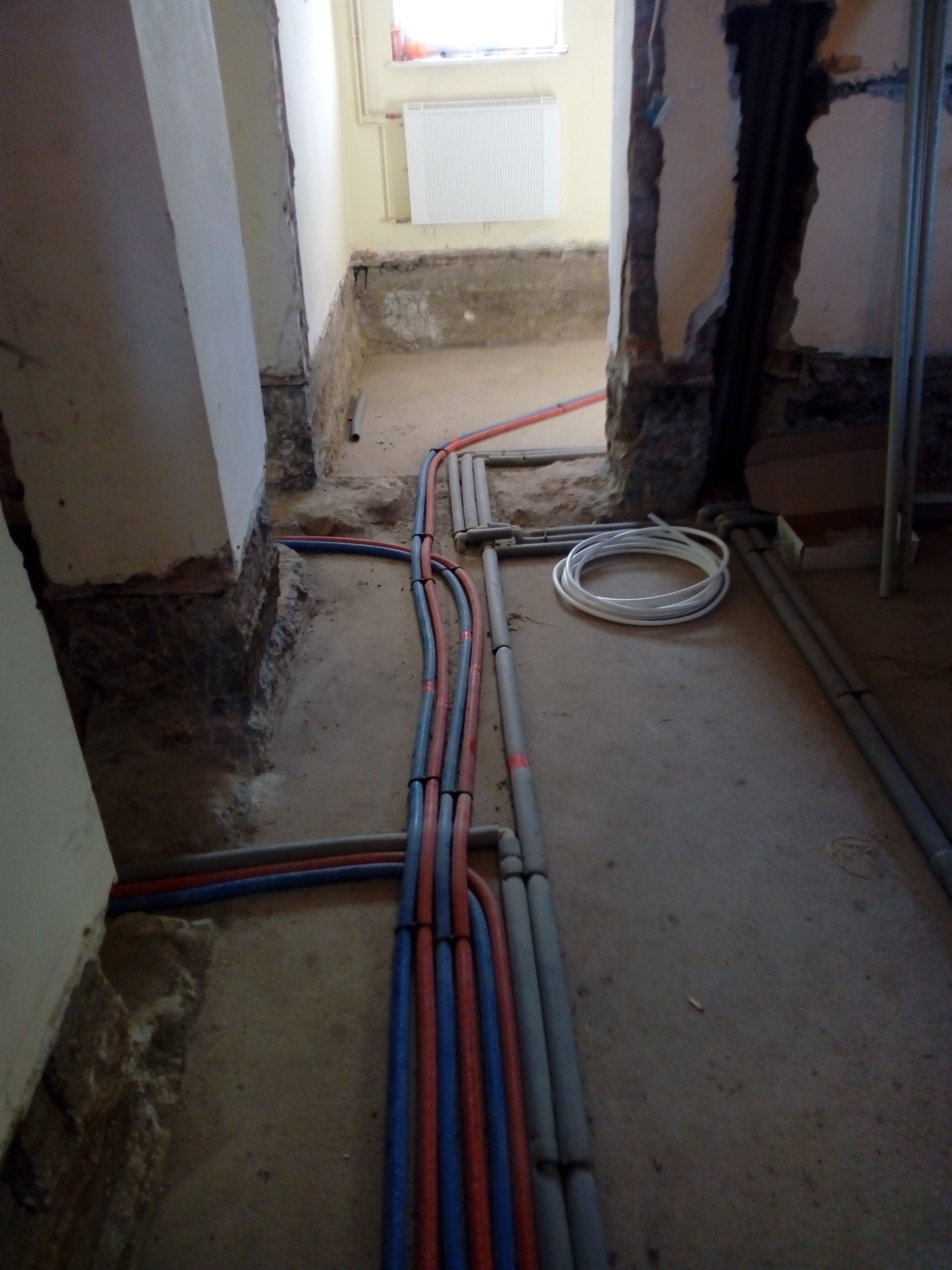 Wnętrze w trakcie remontu z odsłoniętymi instalacjami: rury grzewcze i wodociągowe w kolorach niebieskim, czerwonym i szarym, prowadzone wzdłuż ściany i podłogi. Fragment ściany z odsłoniętą cegłą...