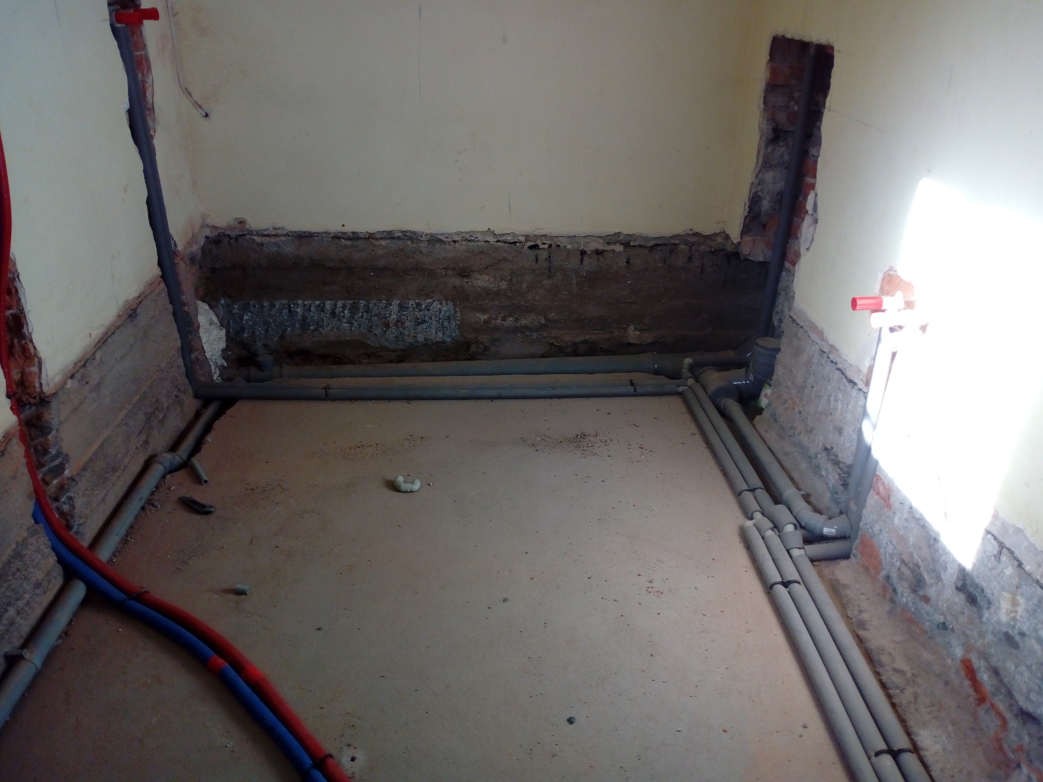 Surowe wnętrze w trakcie hydraulicznych prac instalacyjnych: szare rury ułożone wzdłuż ścian z odsłoniętą cegłą, widoczne wnęki w ścianach przygotowane pod instalacje, czerwono-niebieskie rury...