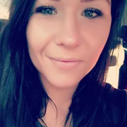 Portret uśmiechniętej brunetki z niebieskimi oczami, skierowanej wprost, z naturalnym makijażem i delikatnym oświetleniem.