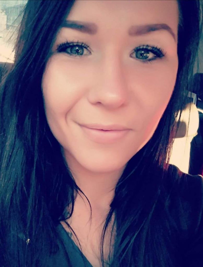 Portret uśmiechniętej brunetki z niebieskimi oczami, skierowanej wprost, z naturalnym makijażem i delikatnym oświetleniem.