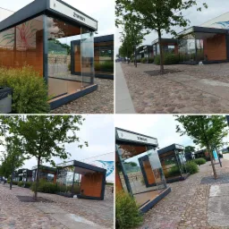 Szereg nowoczesnych, przeszklonych pawilonów z drewnianym wykończeniem, ustawionych wzdłuż brukowanej ulicy, z widocznymi drzewami i muralem w tle.