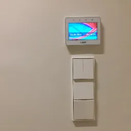 Panel sterowania systemem alarmowym Safel z wyświetlaczem daty i godziny, zamontowany na ścianie obok potrójnego włącznika światła.