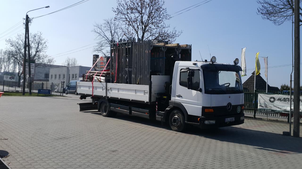 Biała ciężarówka z platformą załadowana metalowymi konstrukcjami rusztowań, zabezpieczonymi czerwonymi pasami, widoczna z boku na brukowanym placu.