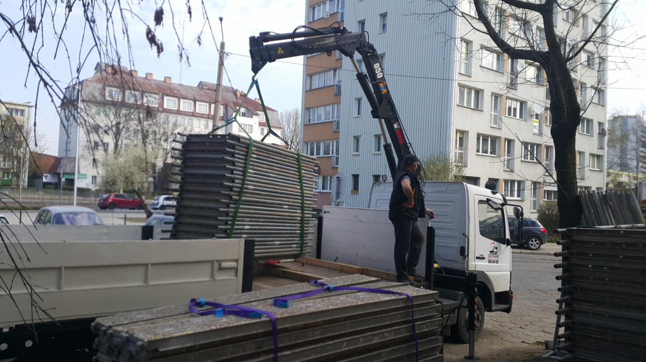 Operator na platformie ciężarówki z HDS podnosi paletę rur na tle miejskiego krajobrazu.