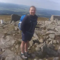 Uśmiechnięta kobieta z niebieskim plecakiem stoi na szczycie góry, otoczona kamieniami i z widokiem na odległe wzgórza.