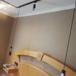 Czarna szyna oświetleniowa zamontowana na suficie nad drewnianym łóżkiem z półką nocną i grawerowanym kogutem na wezgłowiu, w pokoju o beżowych ścianach.