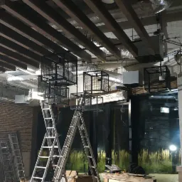 Montaż czarnych, klatkowych lamp wiszących na drewnianym suficie w lokalu, widoczne drabiny, instalacje pod sufitem i ceglana ściana w tle.