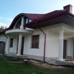 Nowoczesny dom jednorodzinny z białą elewacją, kolumnami i oryginalnym dachem z czerwonej dachówki, widoczne elementy wykończeniowe przy fundamentach