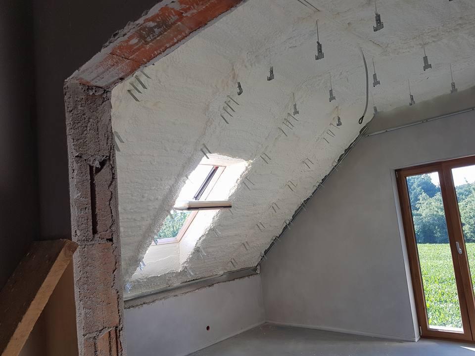Poddasze z oknem dachowym po ociepleniu pianką poliuretanową, widoczne metalowe uchwyty mocujące, surowy mur w tle, okno z widokiem na zieleń.