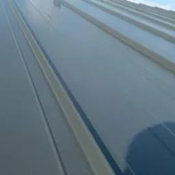 Szary dach z blachy trapezowej z kominkiem wentylacyjnym na tle błękitnego nieba.