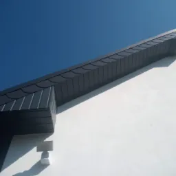 Fragment dachu z szarej blachy, widoczny z dołu na tle błękitnego nieba, z elementami obróbek blacharskich.