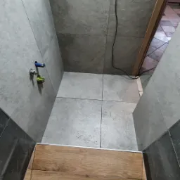 Wnętrze łazienki w trakcie remontu: szare płytki ścienne i podłogowe, fragment drewnopodobnej podłogi, widoczne niedokończone prace, w tym wystające przewody i niezakończone fugowanie, oraz para...