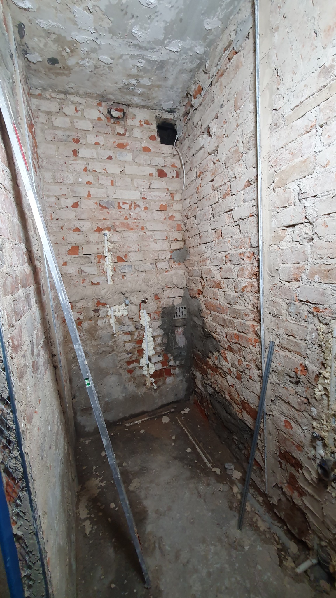 Surowe wnętrze z ceglanymi ścianami w trakcie prac przygotowawczych, widoczne instalacje i poziomica oparta o ścianę.