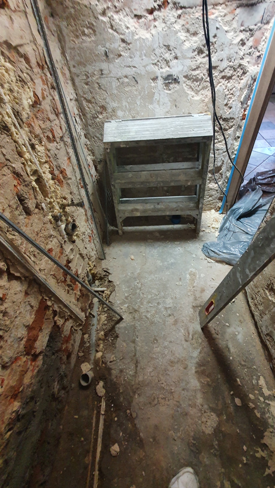 Surowe wnętrze w trakcie prac: odsłonięta cegła, instalacje, prowizoryczna konstrukcja z drewna, widoczne ślady po kuciu i demontażu, podłoga z resztkami gruzu i kurzu.