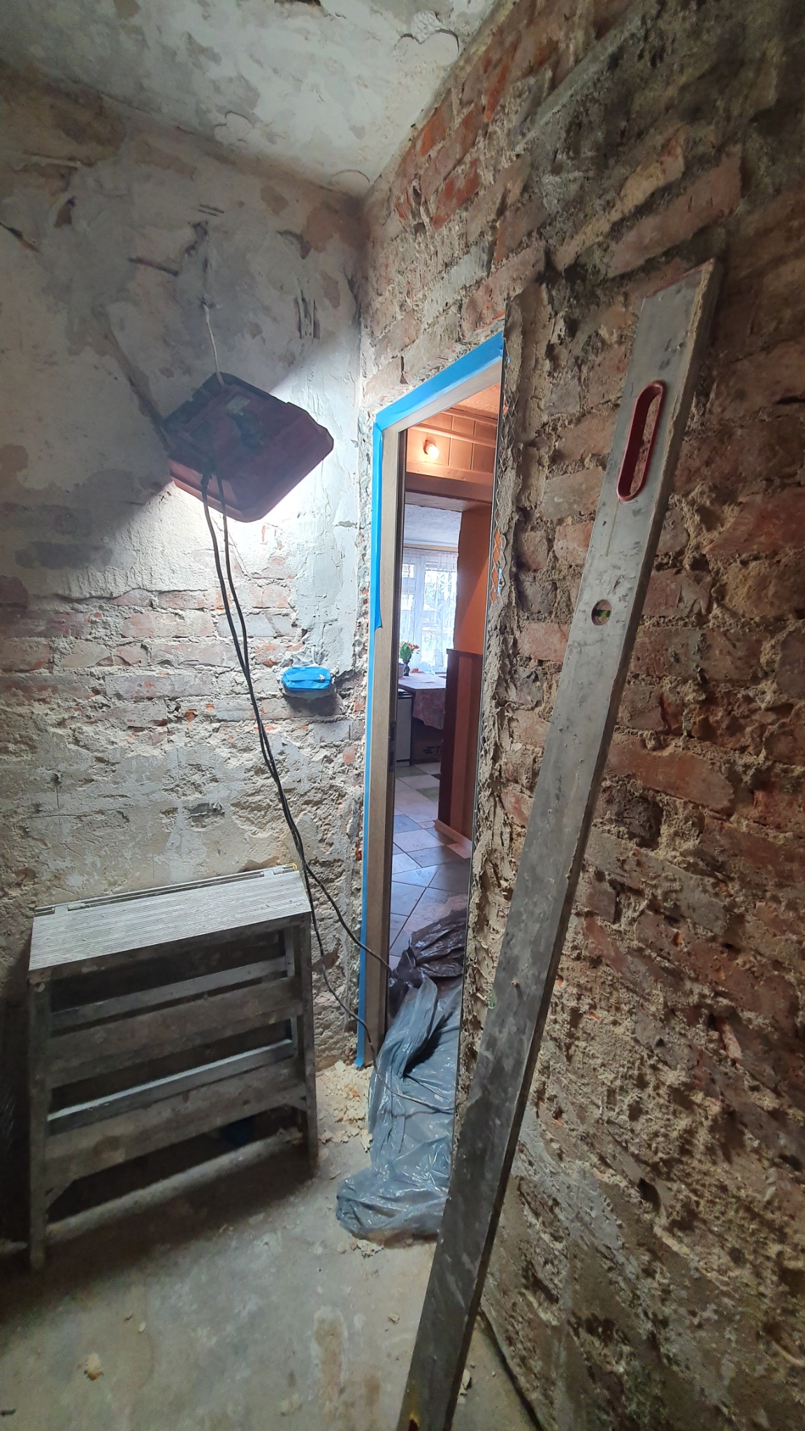 Wnętrze w trakcie prac z odsłoniętą cegłą, zabezpieczonym otworem drzwiowym niebieską folią, widoczna poziomica oparta o ścianę, prowizoryczne oświetlenie i drabinka.