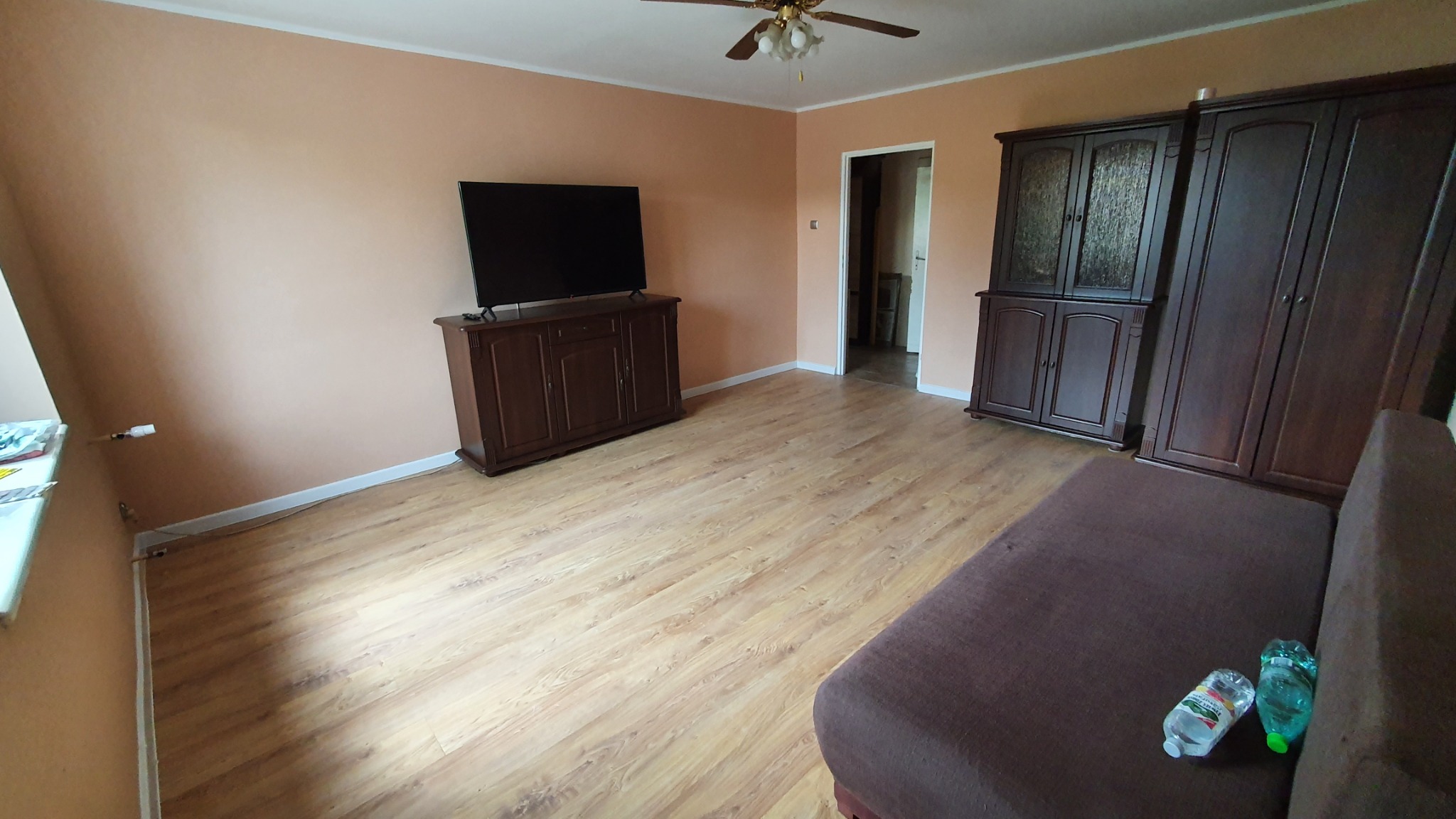 Salon z drewnianą podłogą, brązowymi meblami i telewizorem na komodzie, ściany pomalowane na brzoskwiniowo, widoczne butelki z wodą na kanapie.