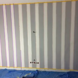 Przygotowana ściana do malowania w pionowe pasy, z taśmą malarską wyznaczającą granice, widoczne narzędzia na folii ochronnej.