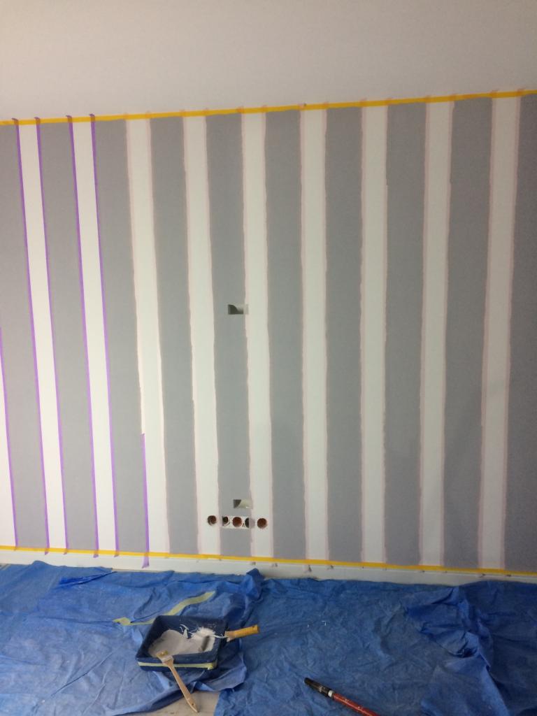 Przygotowana ściana do malowania w pionowe pasy, z taśmą malarską wyznaczającą granice, widoczne narzędzia na folii ochronnej.