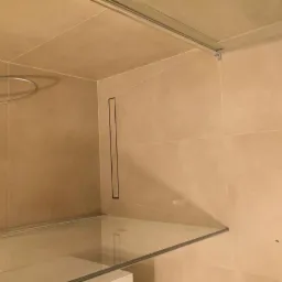 Beżowe płytki w łazience z odpływem liniowym w ścianie, widoczna szklana kabina prysznicowa i słuchawka prysznicowa.