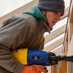 Pracownik w pomarańczowych rękawicach montuje płytę warstwową przy użyciu niebiesko-ż&oacute;łtej gwoździarki B.PRO, widoczna konstrukcja drewniana i fragment nieba.