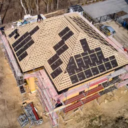 Widok z góry na budowę dachu domu jednorodzinnego; widoczne rusztowania, drewniana konstrukcja łat, czarna membrana dachowa z nadrukiem, dwóch pracowników na dachu oraz częściowo ocieplone ściany...