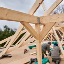 Konstrukcja drewniana dachu w trakcie montażu, widoczne elementy złączne, deski, płyty OSB, piła łańcuchowa i dwóch pracowników w ubraniach roboczych z logo firmy WatraDom.