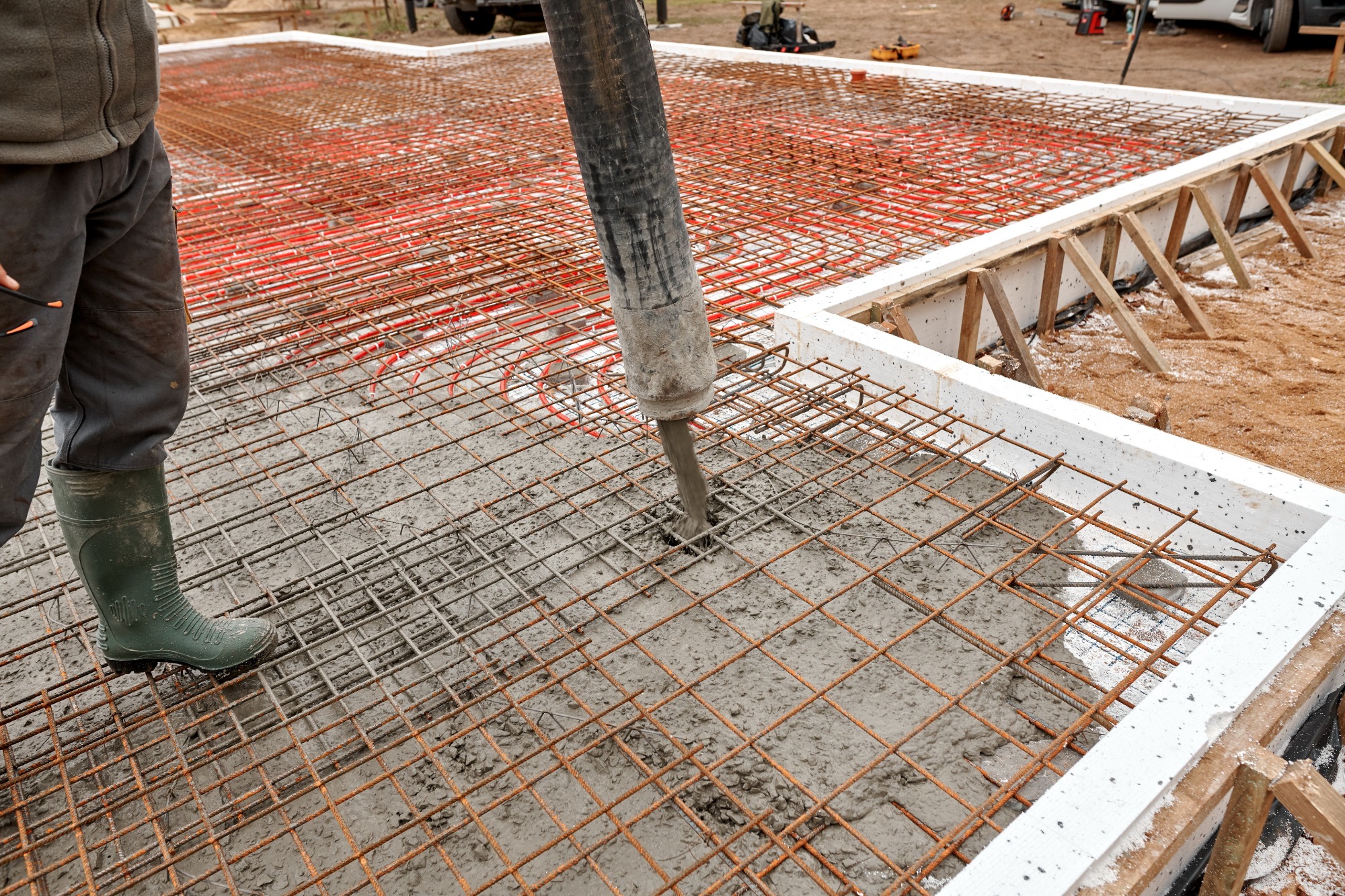Wylewanie betonu na zbrojenie fundamentu z rurami ogrzewania podłogowego, widoczna noga w zielonym gumowcu i wąż pompy do betonu.