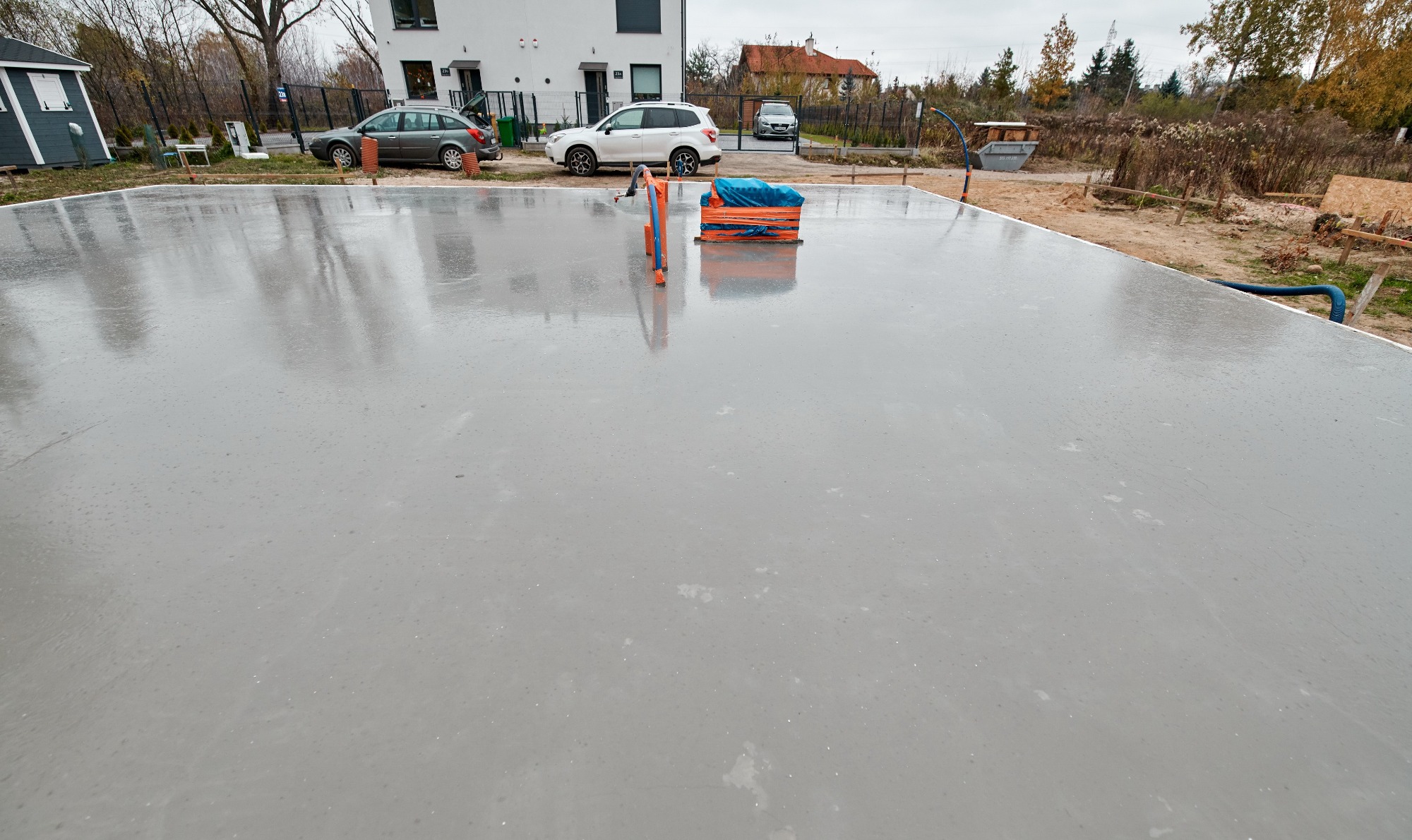 Świeżo wylana, gładka, betonowa płyta fundamentowa, częściowo mokra od deszczu, z widocznymi narzędziami i materiałami budowlanymi na obrzeżach, w tle domy i samochody.