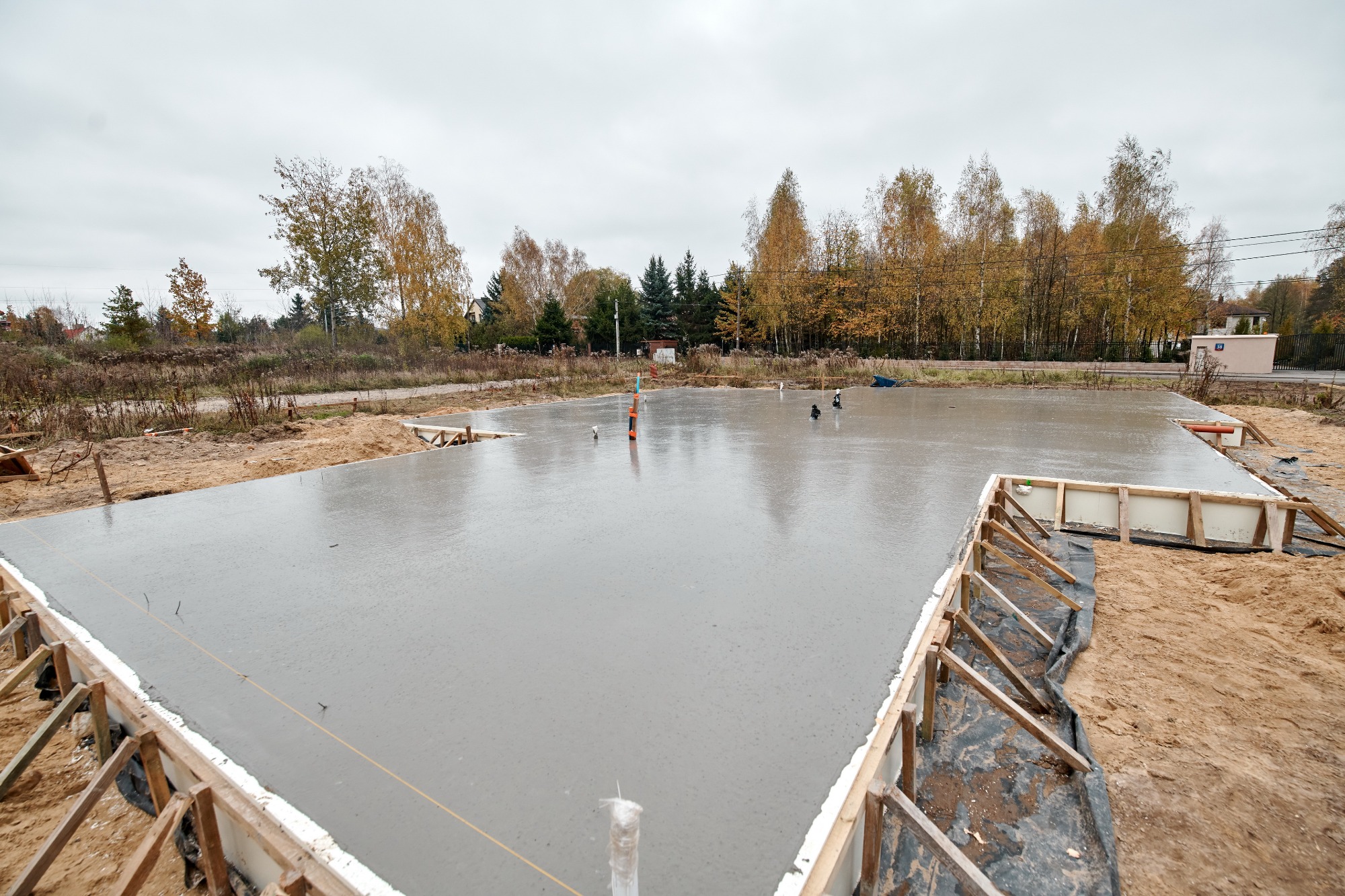 Wylany fundament pod budynek w Łodzi, z widocznym szalunkiem i wystającymi rurami instalacyjnymi na tle jesiennego krajobrazu.