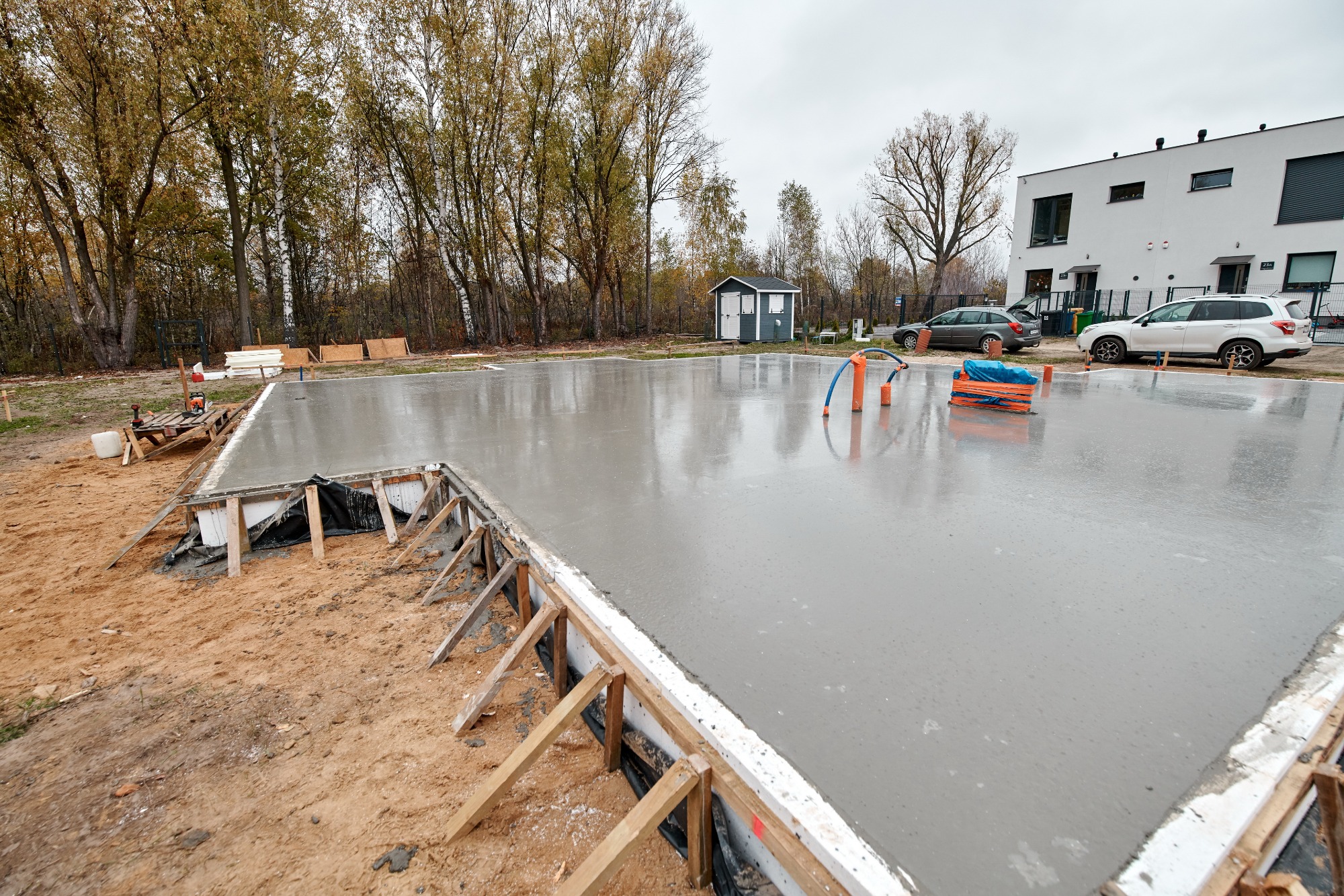 Świeżo wylana, gładka płyta fundamentowa z drewnianym szalunkiem, widoczne pomarańczowe rurki i niebieski wąż na powierzchni betonu, w tle budynek mieszkalny i drzewa.
