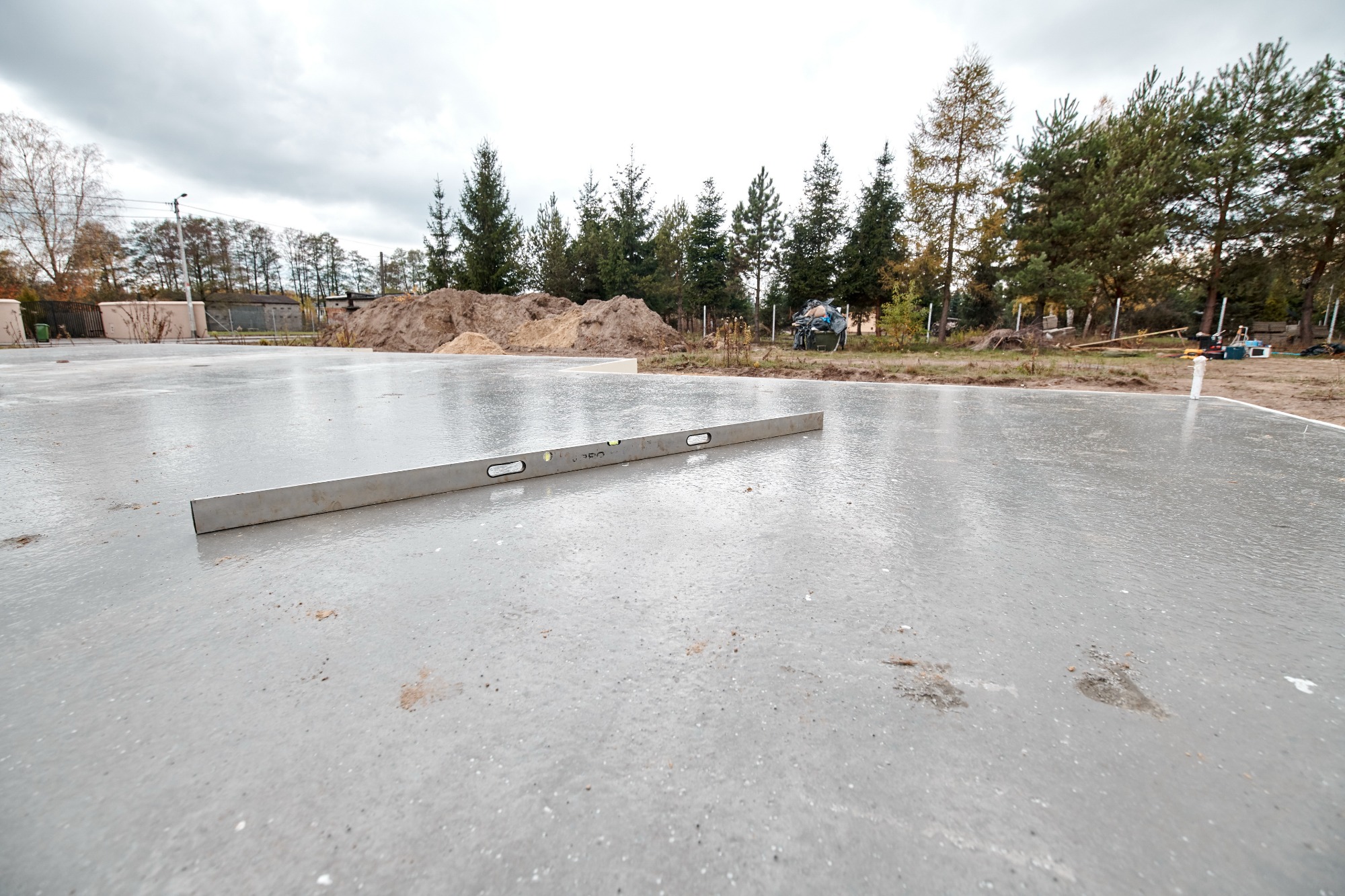 Wylana i wypoziomowana płyta fundamentowa z poziomnicą na powierzchni, widoczne ślady zabrudzeń i wilgoci, w tle hałda ziemi i drzewa.