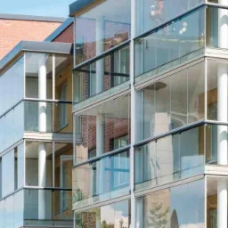 Zewnętrzny widok budynku z balkonami zabudowanymi systemem przesuwnych szklanych paneli z czarnymi ramami i białymi kolumnami, z widoczną ceglaną elewacją i błękitnym niebem w tle.