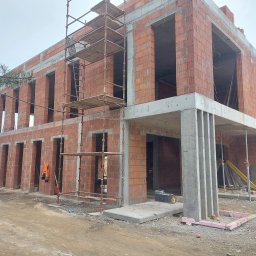 Usługi Budowlane Janusz Stawicki - Dwukondygnacyjny budynek w stanie surowym otwartym, wzniesiony z czerwonej cegły kratówki, z widocznymi szalunkami i rusztowaniami, na placu budowy z ziemistym podłożem.