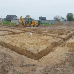 Żółta koparko-ładowarka JCB przy wykopie pod fundamenty domu jednorodzinnego, widoczne zbrojenie w wykopie, teren przygotowany pod wylewkę.