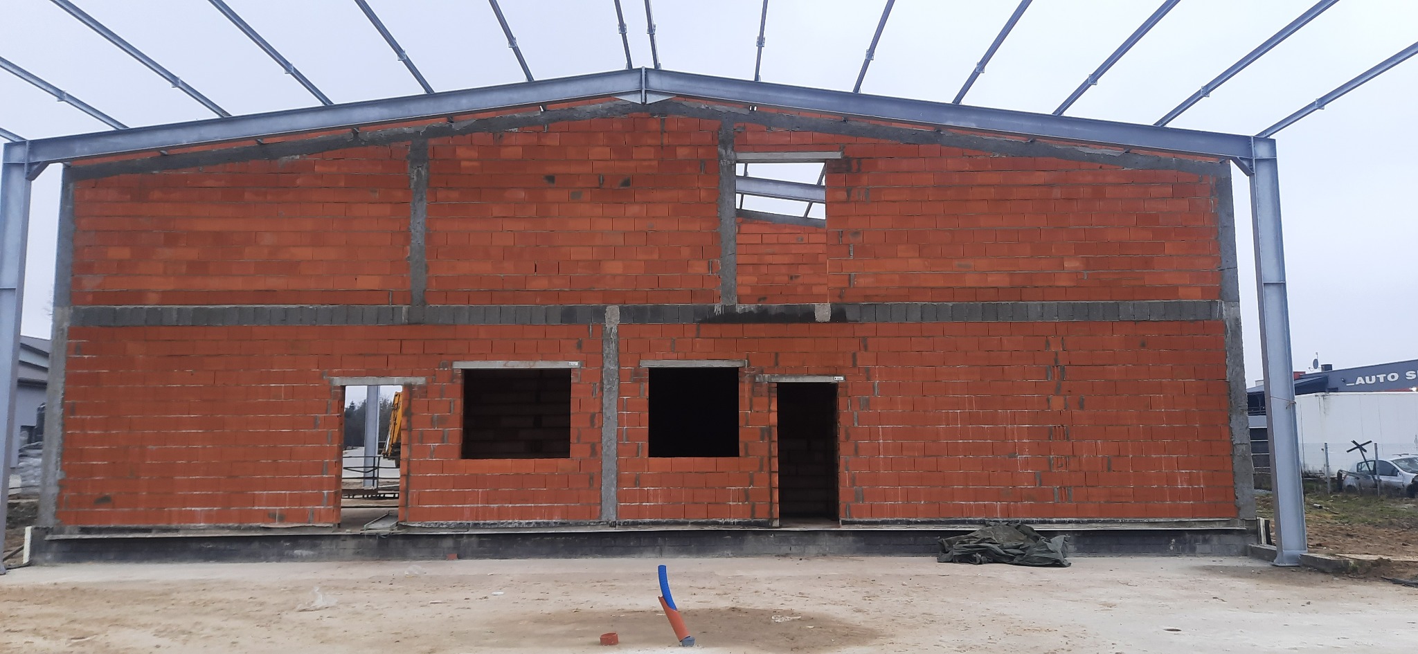 Surowy stan budynku z czerwonej cegły z widoczną stalową konstrukcją dachu i otworami okiennymi oraz drzwiowymi, teren budowy z piaskiem i fragmentem rury PCV
