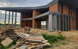 Surowy szkielet domu z czerwonej cegły, widoczne betonowe kolumny i zadaszenie, w tle pole i pochmurne niebo, na pierwszym planie stos desek i betoniarka.