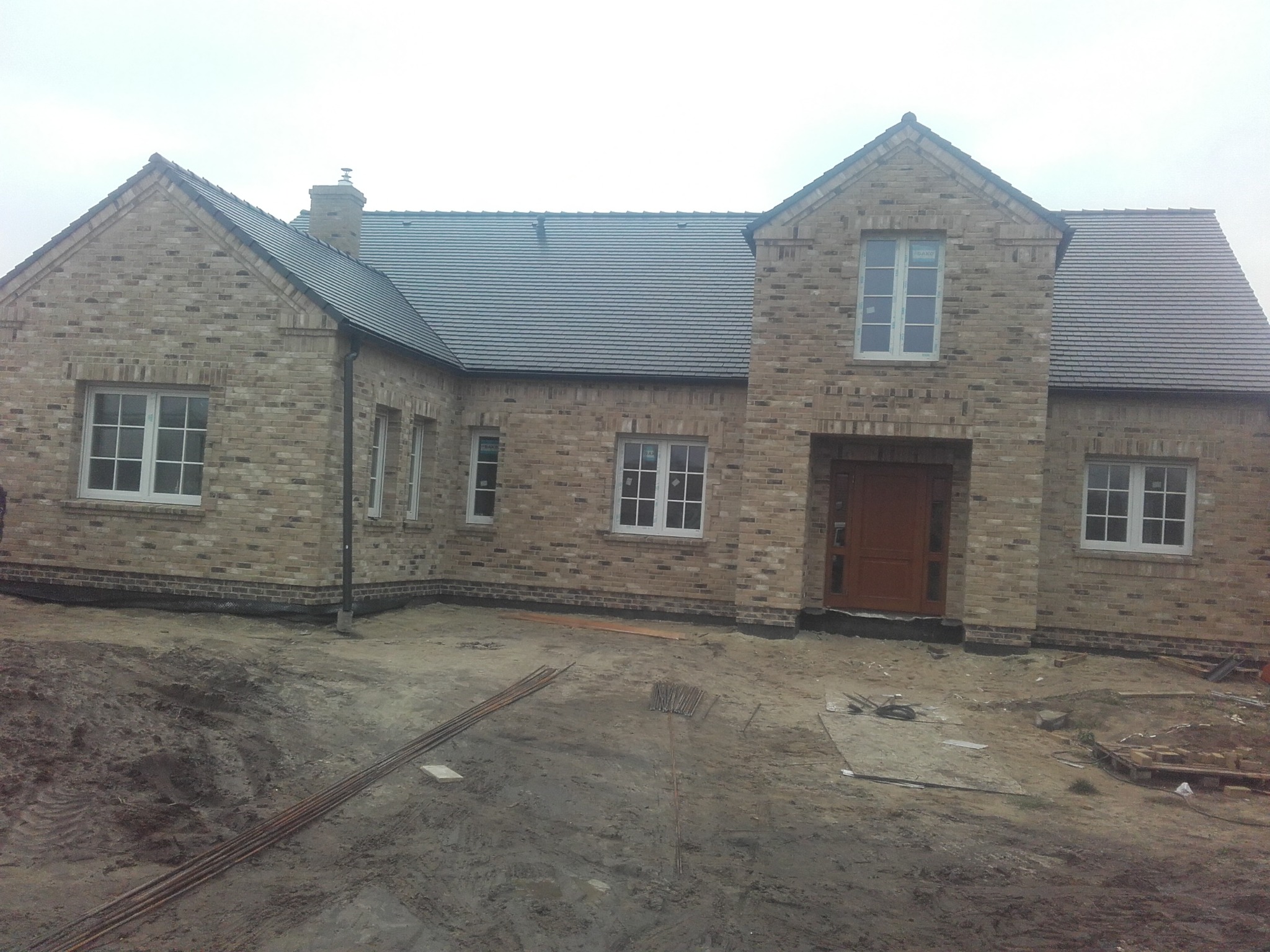 Surowy, dwukondygnacyjny dom z jasnej cegły z ciemnoszarą dachówką, białą stolarką okienną i drewnianymi drzwiami wejściowymi, na etapie wykończenia terenu.