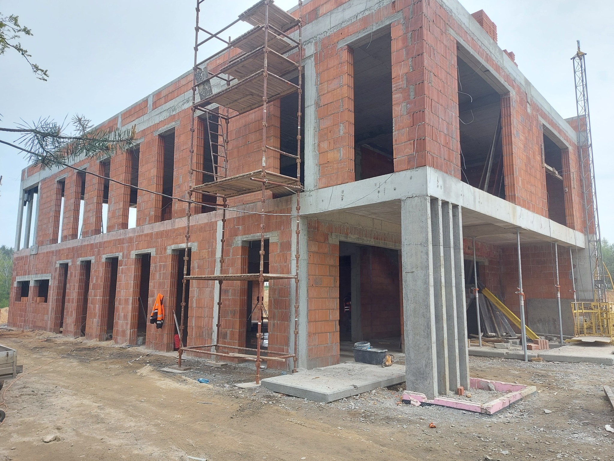 Dwukondygnacyjny budynek w stanie surowym otwartym, wzniesiony z czerwonej cegły kratówki, z widocznymi szalunkami i rusztowaniami, na placu budowy z ziemistym podłożem.