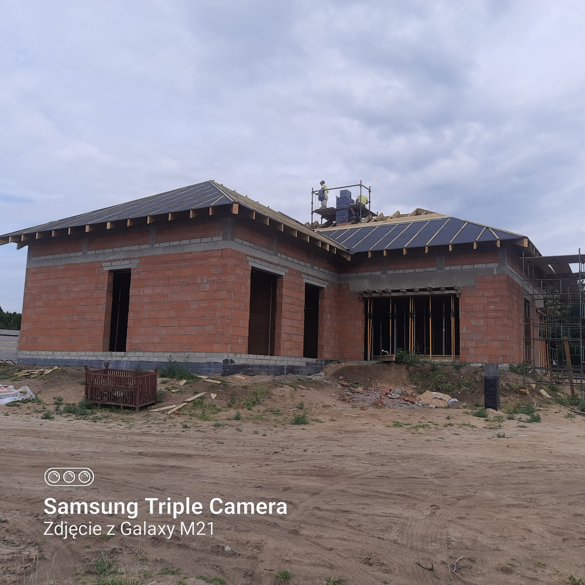 Budowa domu jednorodzinnego z czerwonej cegły, widoczne niezadaszone otwory okienne i drzwiowe, pracownicy na rusztowaniu układają dachówkę, teren wokół w stanie surowym.