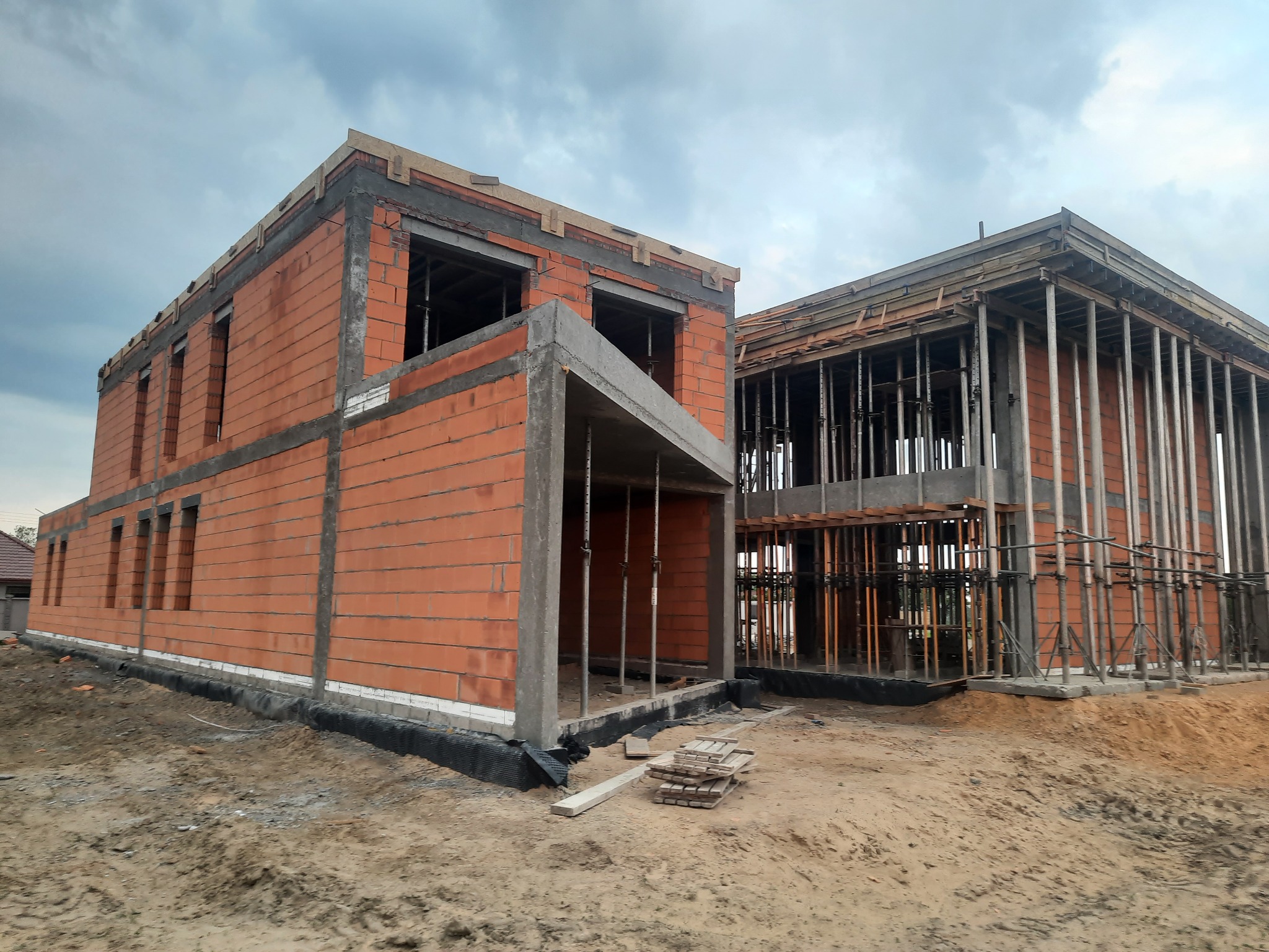Wznoszenie dwukondygnacyjnego budynku z czerwonej cegły ceramicznej, widoczne szalunki stropowe wsparte stalowymi podporami, teren budowy w stanie surowym otwartym.