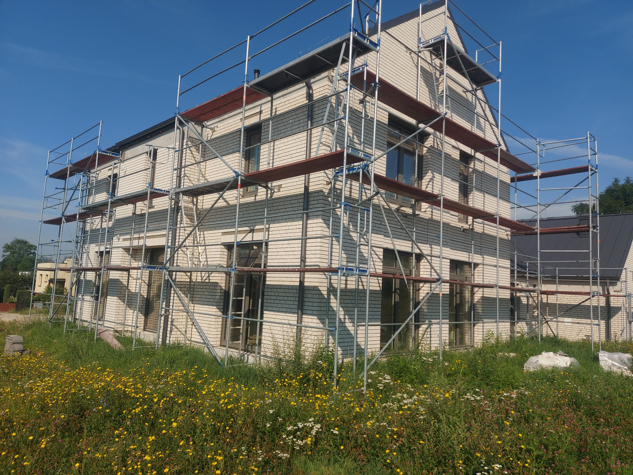 Dwupiętrowy dom murowany w trakcie budowy, otoczony rusztowaniami, z widoczną elewacją z jasnej cegły z ciemniejszymi pasami i bujną roślinnością w otoczeniu.