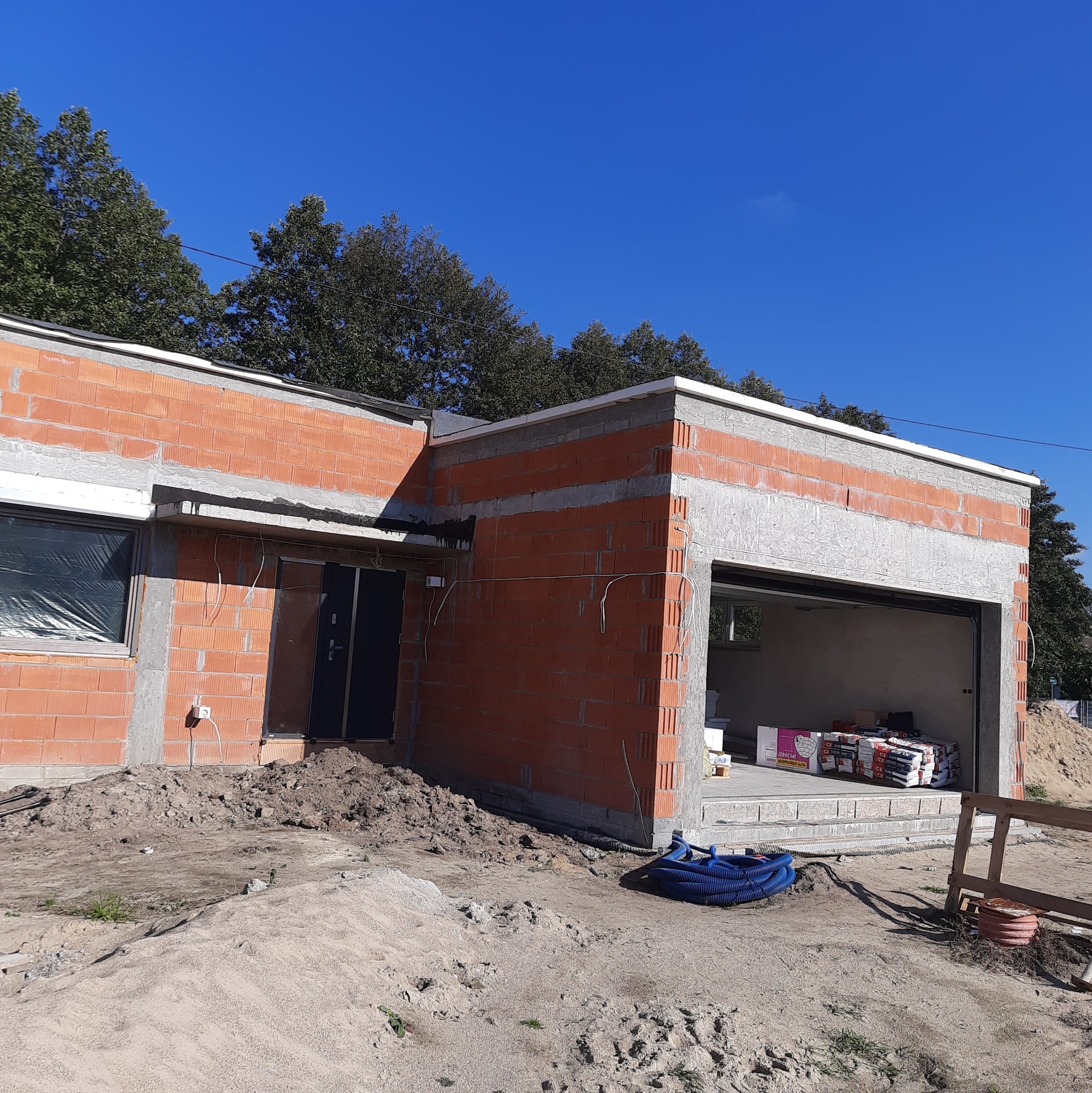 Niedokończony budynek z czerwonej cegły z widocznym otworem garażowym, materiałami budowlanymi w środku i niebieskim przewodem na zewnątrz, w słoneczny dzień.
