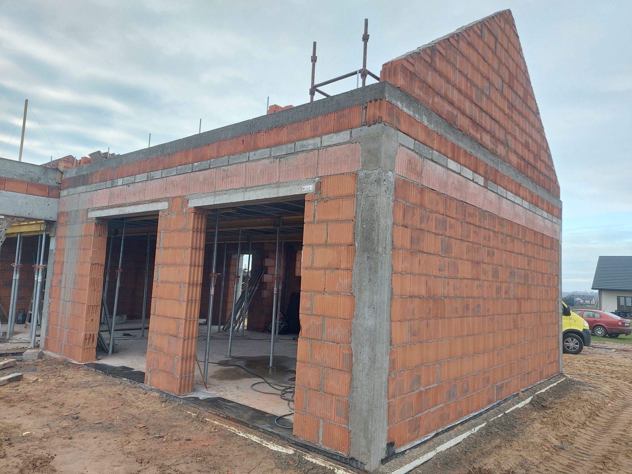 Budowa domu z czerwonej cegły ceramicznej, widoczne zbrojenia i betonowe wylewki, w tle zaparkowane samochody.